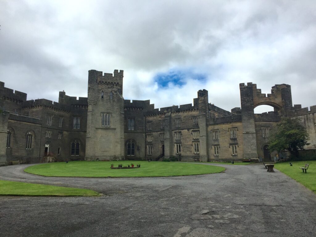A picture of Brancepeth Castle from inside the courtyard. The walls are beige against a mainly cloudy sky with glimpses of blue. The grass is well manicured in the foreground