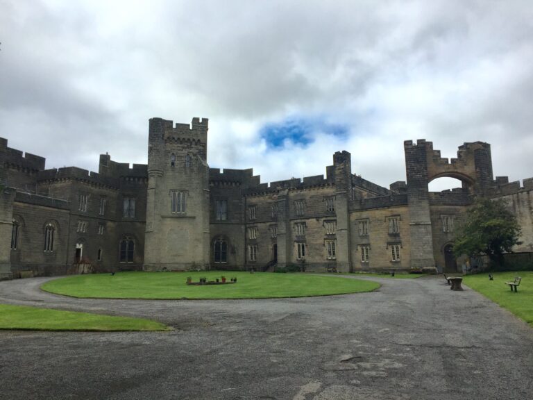 A picture of Brancepeth Castle from inside the courtyard. The walls are beige against a mainly cloudy sky with glimpses of blue. The grass is well manicured in the foreground